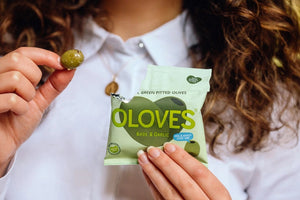 A light green packet of OLOVES - BASIL & GARLIC natural green pitted Greek olives by POSHI, flavored with basil and garlic. The packaging showcases a cartoonish olive graphic and emphasizes: "Low Calorie Snack," "New Greek Olives," "Kids & Adults Love 'Em," "Gluten-Free Vegan Snacks," and "30g | 1.1oz.