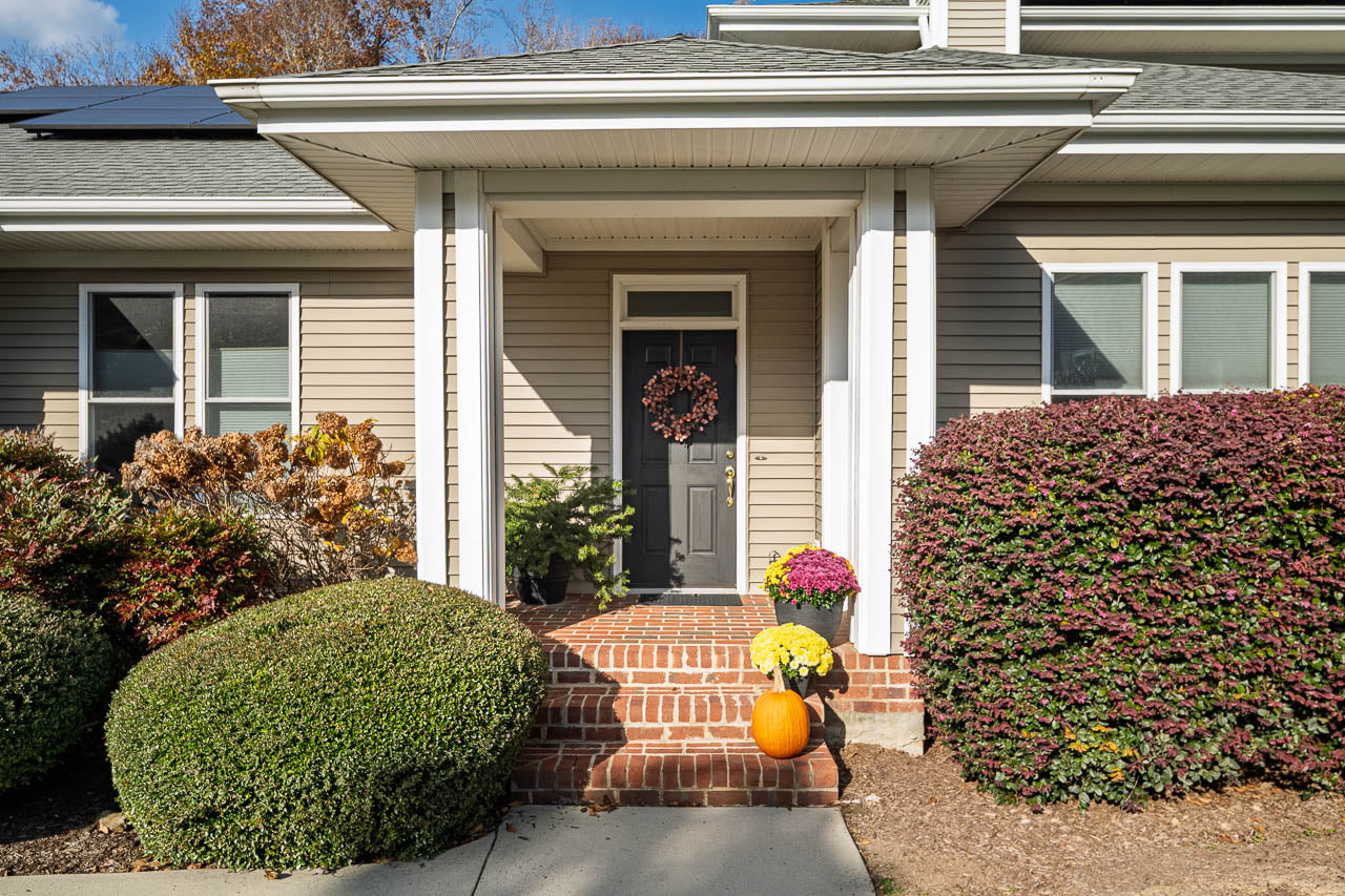 A house entrance with a gray front door decorated with a wreath, brick steps, potted yellow and white flowers, a pumpkin, and neatly trimmed bushes on either side.