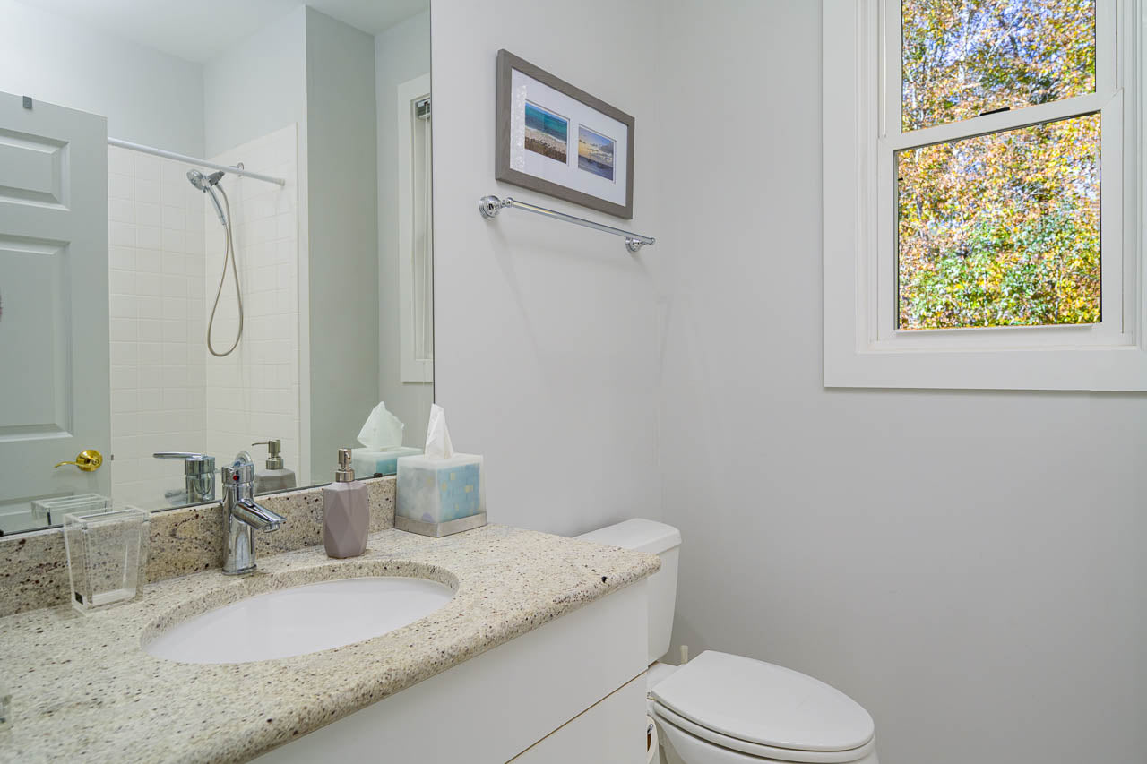 A clean, modern bathroom with a granite countertop, sink, and faucet, a tissue box, soap dispenser, and glass on the vanity, a toilet, and a window showing green trees outside. A beach photo hangs above the towel bar.