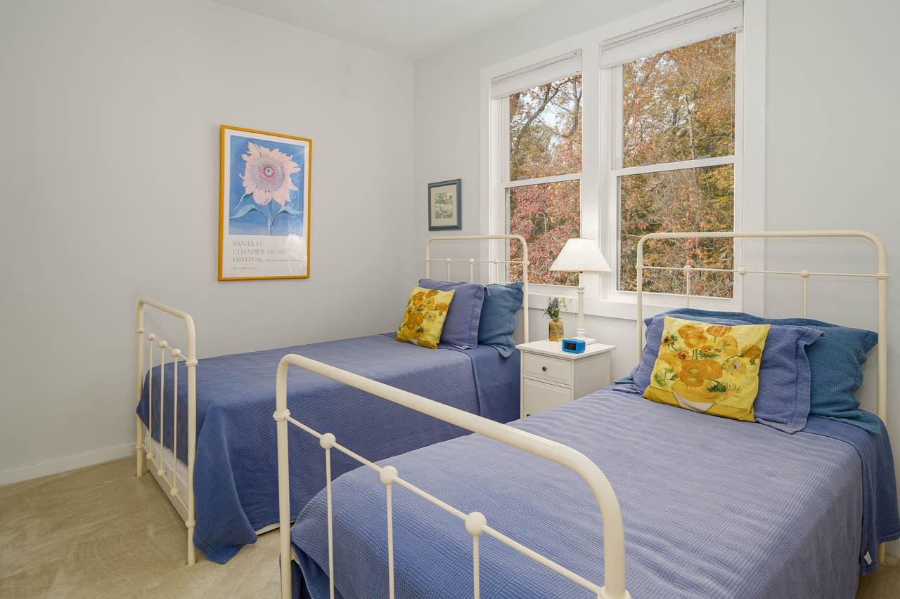 A bright bedroom with two white metal twin beds, blue bedding, yellow floral pillows, a white nightstand with a lamp, and large windows showing autumn trees outside. Artwork decorates the white walls.