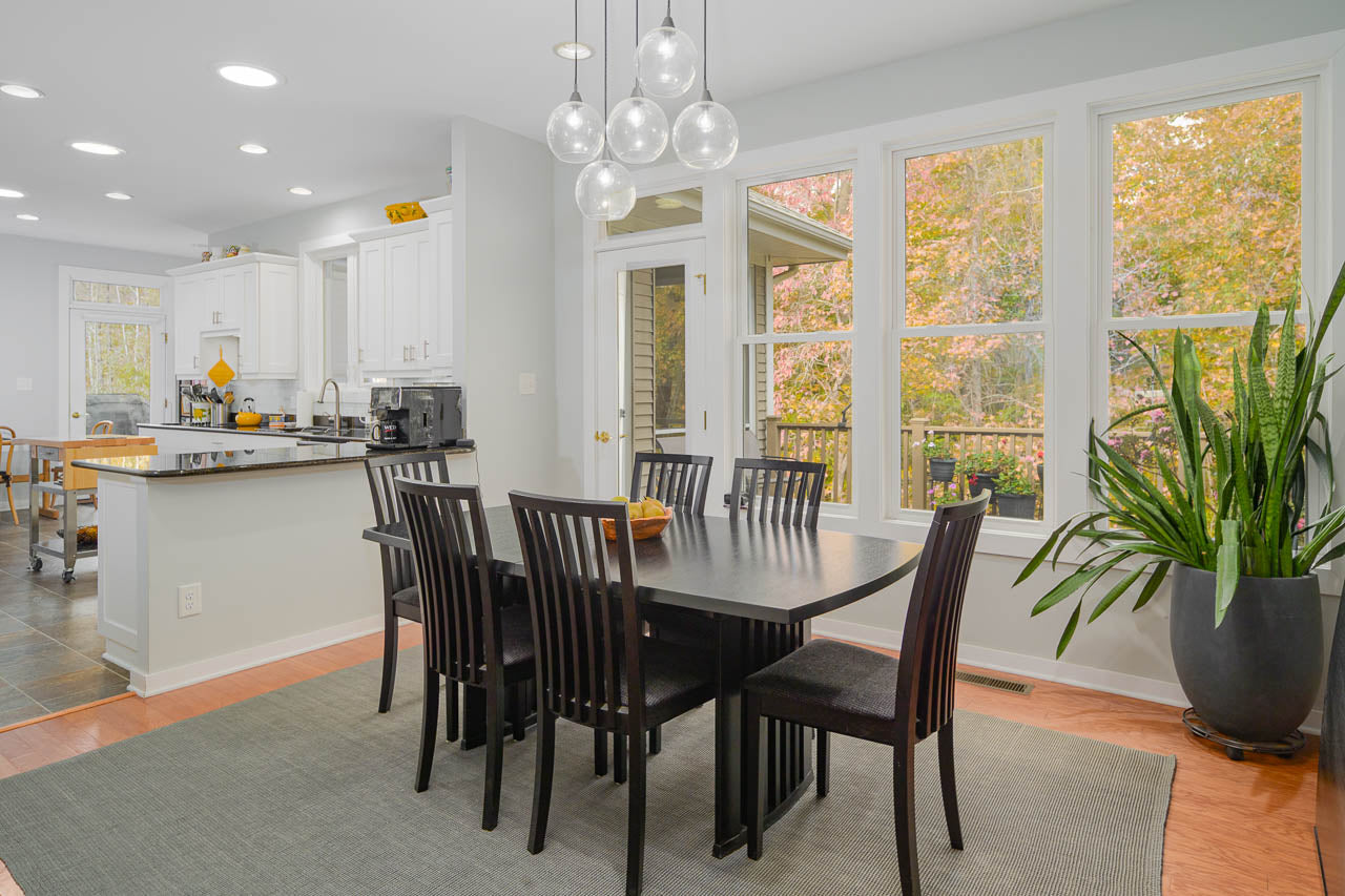 A modern dining room with a black table and six chairs, large windows showing autumn trees, a potted plant, and a kitchen area with white cabinets in the background. Spherical pendant lights hang above the table.