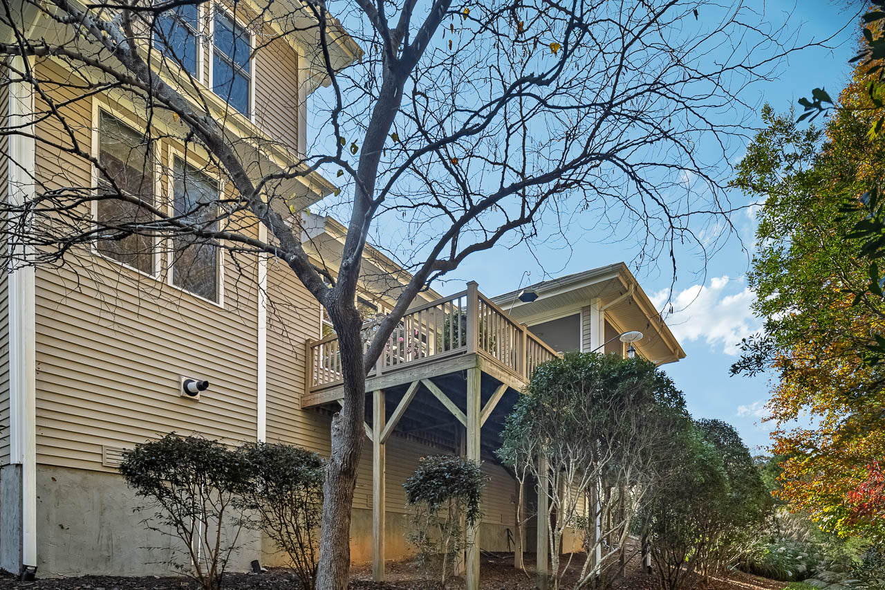 A tan house with a wooden deck, surrounded by leafless trees and shrubs, is shown on a sunny day with a blue sky and scattered clouds.