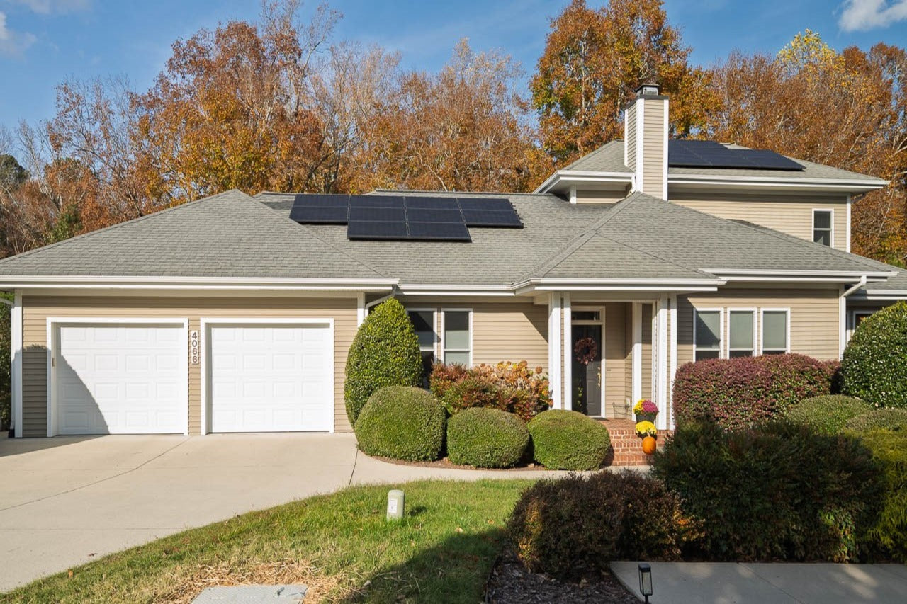 A beige suburban house with two white garage doors, solar panels on the roof, a front porch with potted plants, and neatly trimmed bushes, surrounded by trees with autumn foliage.