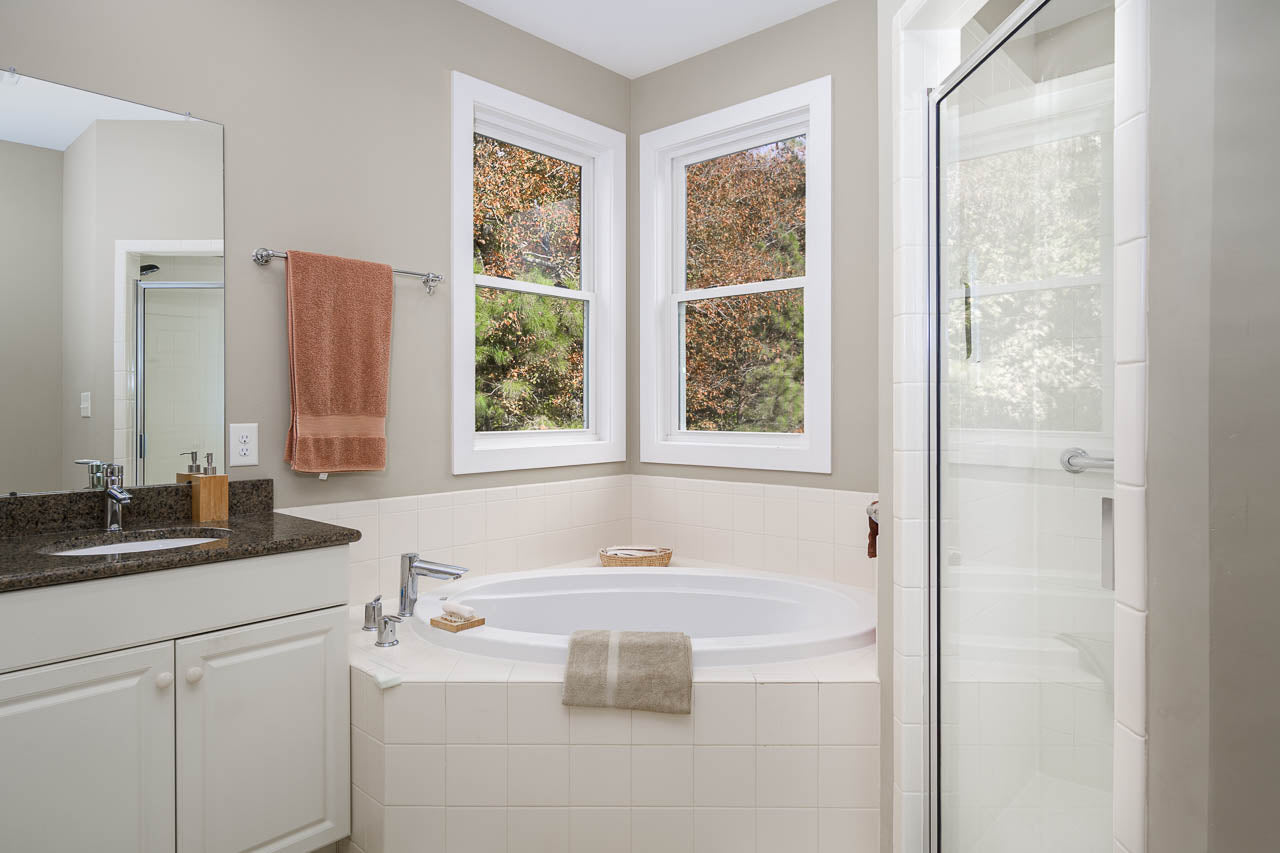Modern bathroom with a large corner bathtub, glass-enclosed shower, white tile walls, two windows with a view of trees, a brown towel hanging on the wall, and a white vanity with a granite countertop.