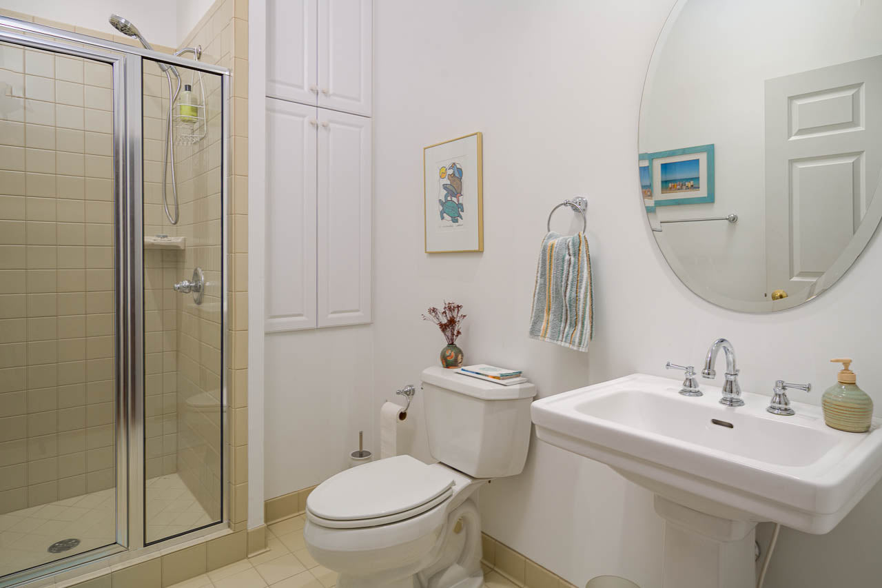 A clean, light-colored bathroom featuring a glass shower, a toilet with books on top, a white pedestal sink with a mirror above, a towel on a ring, and wall art. The floor and shower tiles are beige.