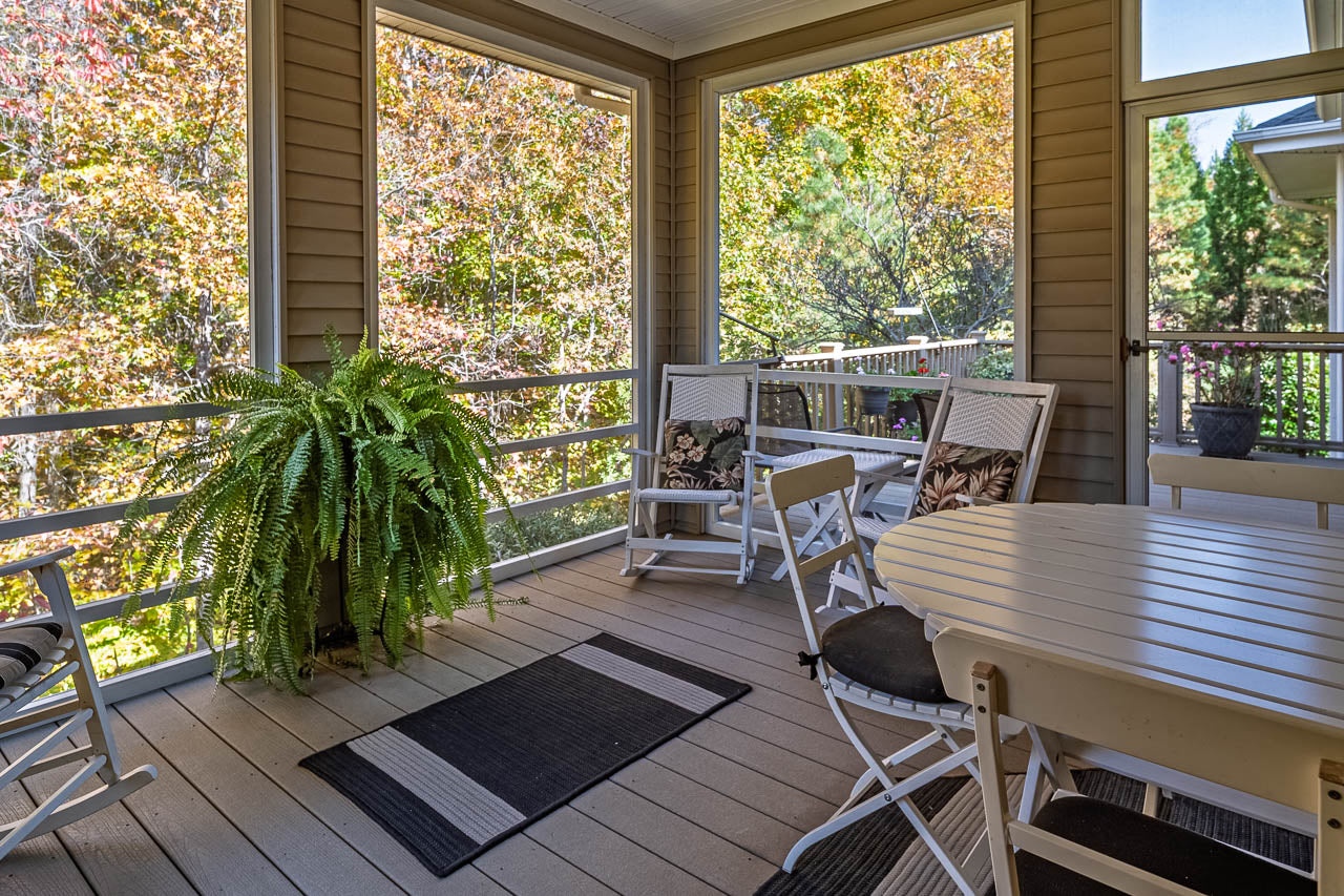 A cozy screened porch features a white table with chairs, rocking chairs, a large green fern, and outdoor rugs. Sunlight filters through the screens, revealing colorful autumn foliage outside.