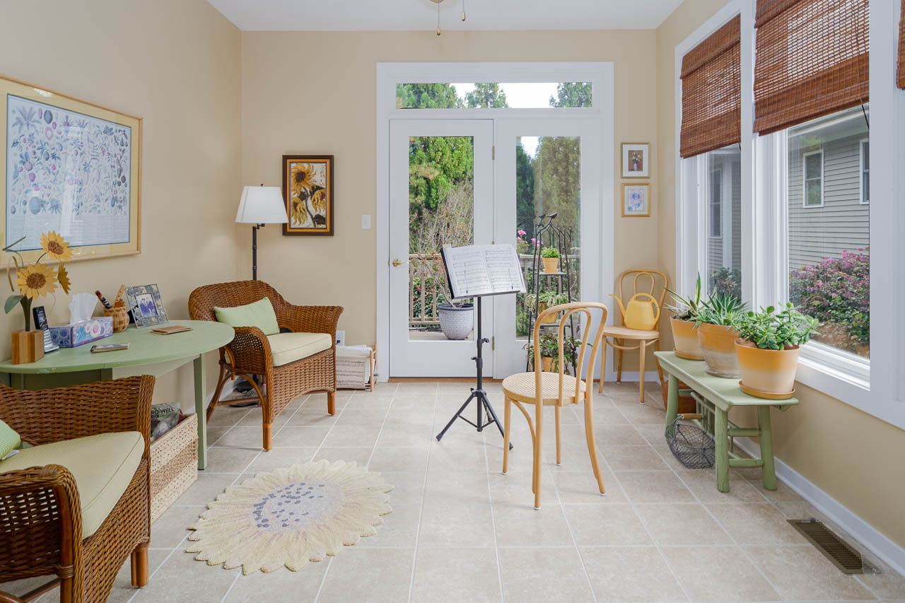 A bright sunroom with wicker furniture, green accents, potted plants, large windows, and a music stand at the center. Light pours in, creating a cozy, inviting atmosphere with a view of greenery outside.