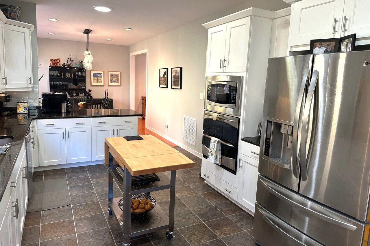 Modern kitchen with white cabinets, stainless steel appliances, a center island with a wooden top, dark countertops, and tiled floor. Framed photos and decorative items are visible on counters and walls.