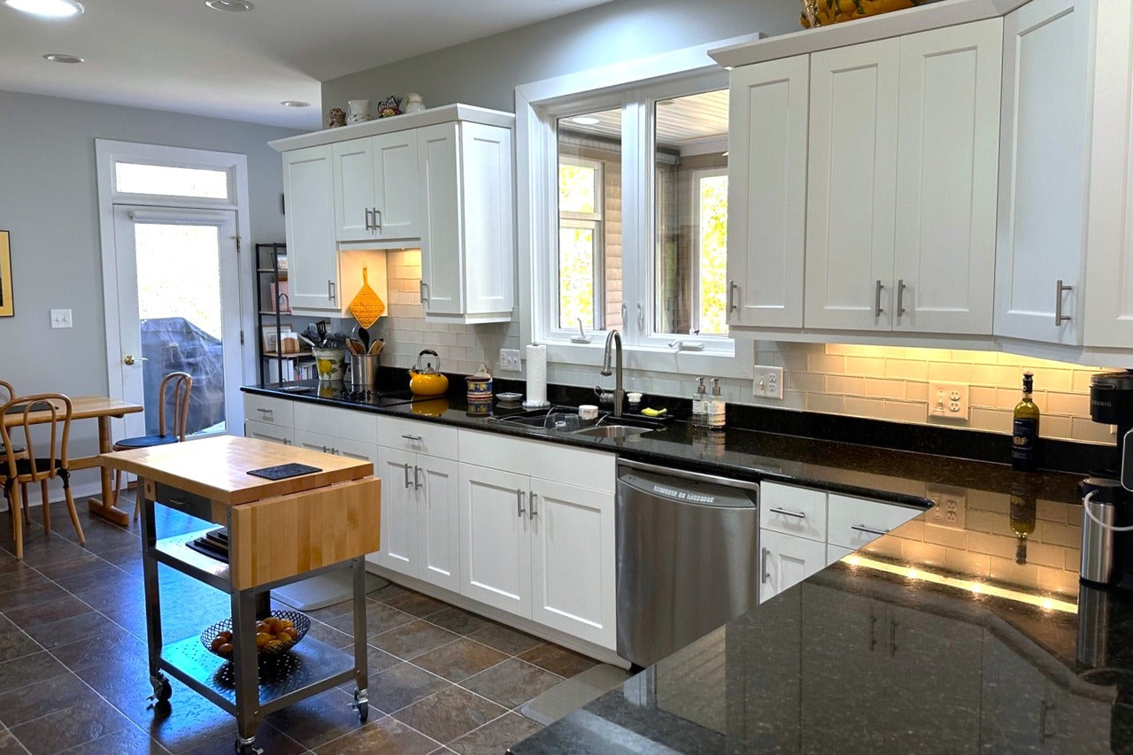 Bright, modern kitchen with white cabinets, stainless steel appliances, a black countertop, and a rolling wooden island. Large window above the sink and a dining area with a wooden table are visible in the background.