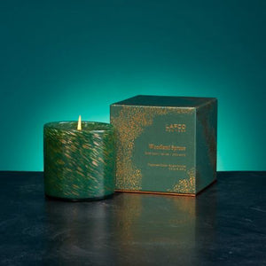 A lit soy wax candle with a green marbled design is positioned beside its coordinating box that reads "LAFCO - WOODLAND SPRUCE HOLIDAY CANDLE 15.5OZ." Housed in a hand-blown glass vessel, it emits the scent of a winter forest. The backdrop features a gradient of teal, enhancing the peaceful atmosphere.