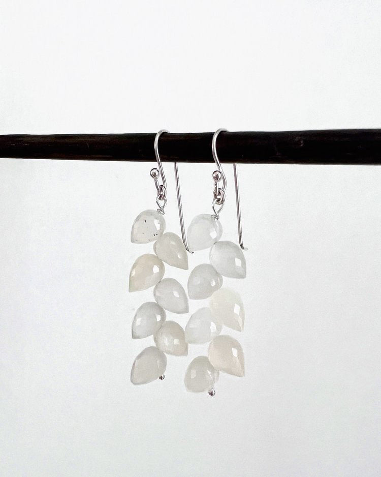 A pair of delicate ANN LIGHTFOOT - LEAF PETAL WHITE MOONSTONE EARRINGS is hanging from a dark rod. Each earring features multiple small, white moonstone teardrop-shaped beads arranged in a cascading design, attached to polished sterling silver earwires. The background is plain white, highlighting the earrings' elegance.