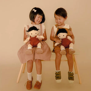 A DINKUM DOLL from OLLI ELLA USA features soft brown hair and comes dressed in a white knit romper paired with brown shoes. This charming doll has an embroidered face with brown eyes, a small nose, and a gentle smile. Its posable body makes it perfect for imaginative playtime, and it is posed standing upright.