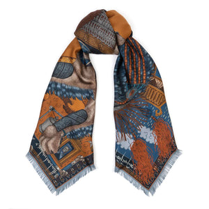 The SABINA SAVAGE - THE SNOW LION SILK TWILL SCARF 135CM by SABINA SAVAGE ENGLAND is a luxurious silk scarf featuring an intricate design in rich browns, blues, and oranges. The pattern, inspired by Tibetan thangkas, includes ornate architectural elements, flowing red hair, and winged figures, all accented by fine detailing and a fringed edge.