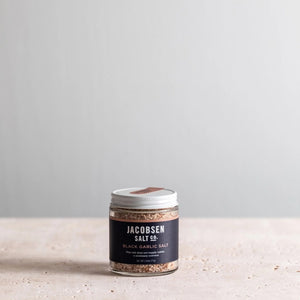 A small glass jar with a white lid, labeled "Jacobsen Salt Co. - Infused Black Garlic Salt," is placed on a light beige surface against a plain, light gray background. The jar contains an aromatic blend of coarse, brownish salt with rich garlic flavor.