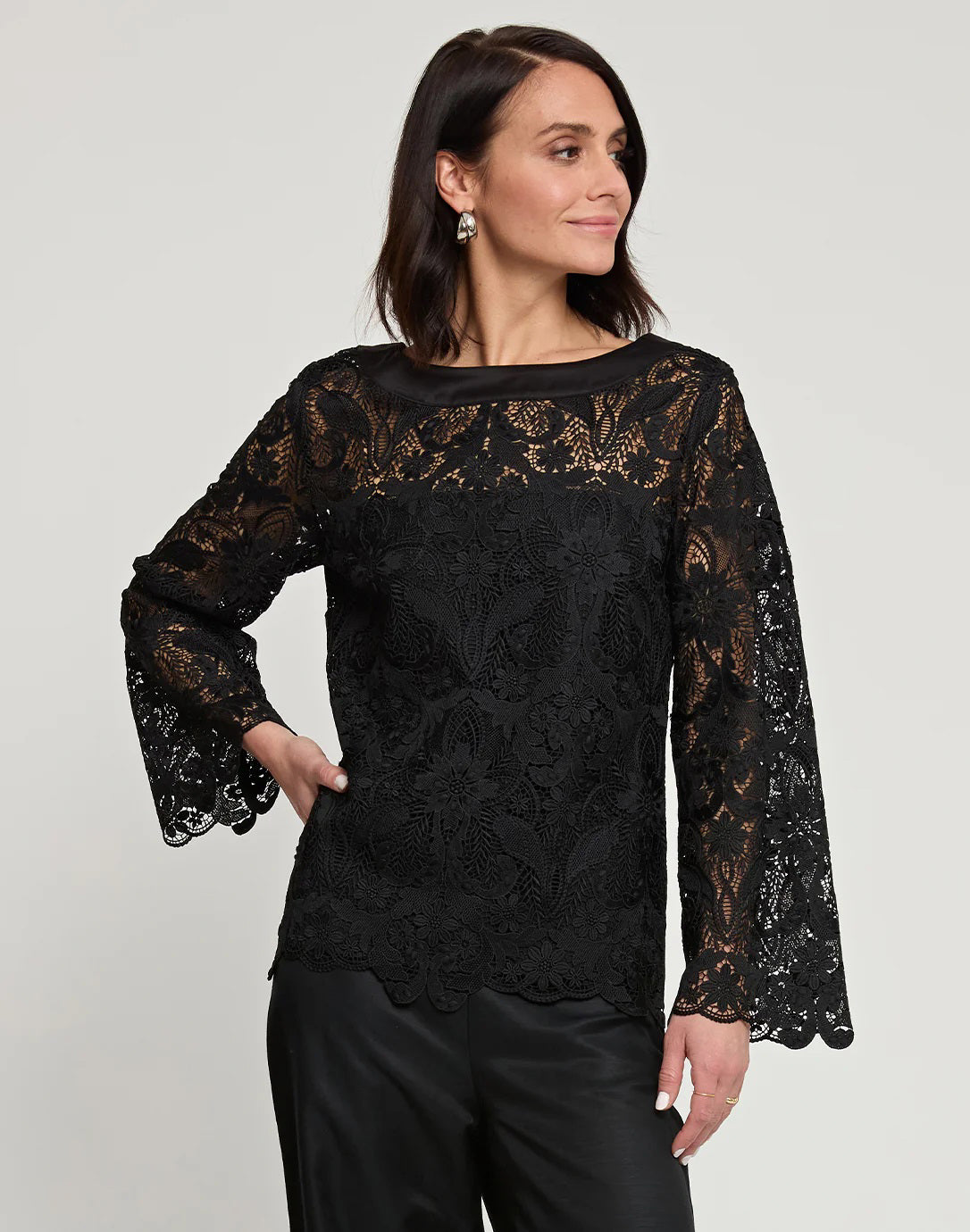 A woman with shoulder-length dark hair is wearing the HINSON WU - BLAKELY LACE TOP, a long-sleeve black lace top that features flared sleeves, paired with black pants. She stands against a plain light gray background, neutrally expressing herself with one hand on her hip.