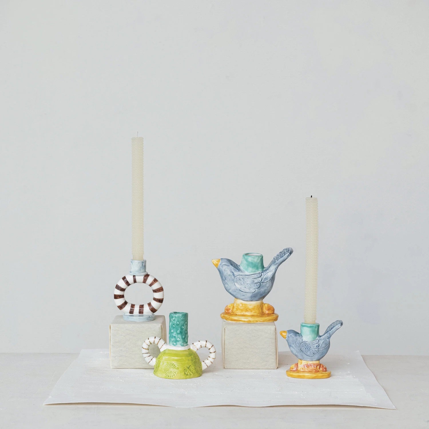 Three minimalist CREATIVE COOP candlesticks display candles; two are HAND PAINTED STONEWARE BIRD TAPER HOLDERS with a navy reactive glaze, and the third features a circular design. Each is uniquely positioned on white blocks against a plain background.