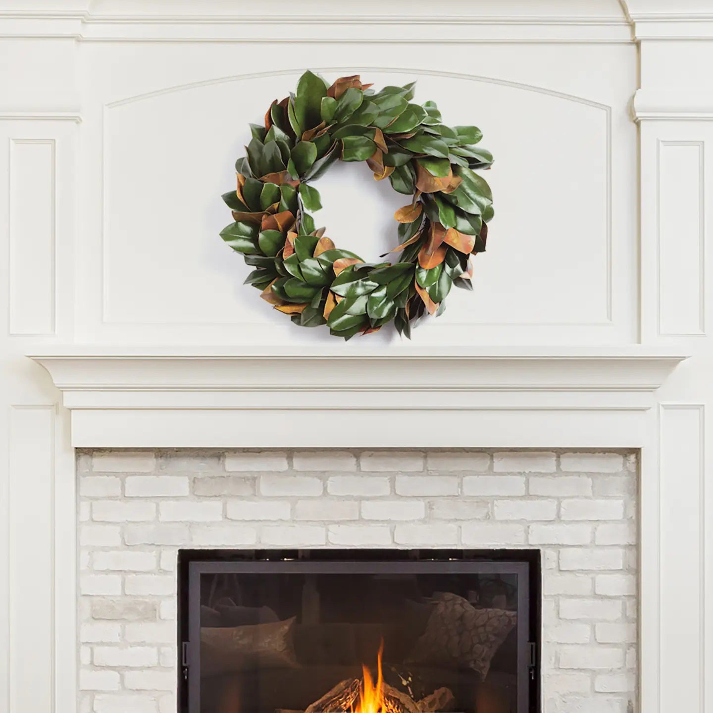 A lush green FAUX MAGNOLIA WREATH 24" from NAPA HOME AND GARDEN adorns a white, ornate fireplace mantel set against a brick surround. Below, a gentle fire burns inside the fireplace, adding warmth to the room's elegant and cozy atmosphere—perfect for interior use as a captivating centerpiece.