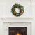 A lush green FAUX MAGNOLIA WREATH 24" from NAPA HOME AND GARDEN adorns a white, ornate fireplace mantel set against a brick surround. Below, a gentle fire burns inside the fireplace, adding warmth to the room's elegant and cozy atmosphere—perfect for interior use as a captivating centerpiece.