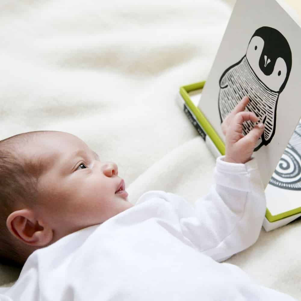 A baby dressed in a white outfit lies on a soft, cream-colored blanket, reaching towards a black-and-white drawing of a penguin on one of the WEE GALLERY - ART CARDS FOR BABIES BLACK AND WHITE. The baby appears to be interacting with the card from WEE GALLERY's set designed to boost cognitive development.