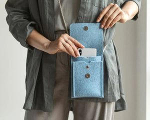A person in a beige sweater and tan pants holds the WOOLBERRY WOOL FELT PHONE BAG, a small gray handcrafted bag with a flap and button closure. The image is cropped at the shoulders and knees.