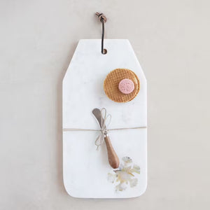 The CHEESE BOARD WITH CANAPE KNIFE by CREATIVE COOP is a rectangular white marble cheese board embellished with a leather hanging loop. It is elegantly decorated with a tied rustic wooden spoon, decorative leaves at the bottom corner, and features a stunning mother of pearl inlay that adds an exquisite touch to any kitchen or dining setting.