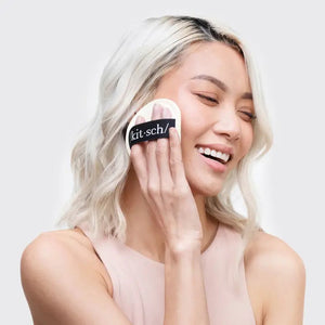 A woman with blonde, wavy hair is smiling and using a round makeup sponge labeled "KITSCH" from her ECO FRIENDLY ULTIMATE CLEANSING KIT to apply product to her face. She is wearing a light pink sleeveless top against a plain white background.