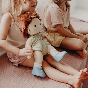 A close-up of a baby wearing OLLI ELLA USA DINKUM DOLL SHOES in Mallow Pink and white socks. The baby is seated on a soft, pale pink textured blanket, dressed in light brown clothing. Only the lower half of the baby's body is visible.