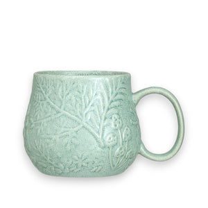 A textured green ceramic teapot and the EMBOSSED BOTANICALS MUG by CREATIVE COOP, both with ornate embossed patterns, sit on a stack of books. The mug is filled with tea, with a teabag nearby. The setting is on a light-colored surface, showcasing the sage reactive glaze beautifully.