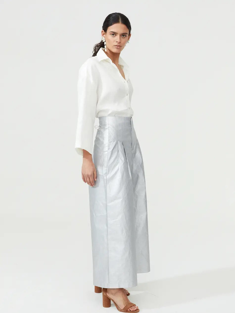 A woman poses against a simple backdrop, dressed in a white blouse with an open collar and high-waisted, wide-leg metallic culotte pants by Lanhtropy. Her hair is tied back and she completes the look with brown open-toed heels.