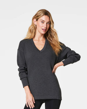 A person with curly hair is standing and looking to the side, wearing a cozy SPANX - AIRESSENTIALS BRUSHED V-NECK TUNIC paired with brown pants. The breathable top drapes elegantly, capturing a sense of laid-back comfort against the plain, light-colored background.