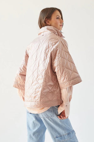 A person with shoulder-length hair stands facing sideways, wearing the ADROIT ATELIER - RUBY QUILTED CAPE, a light pink, short-sleeved jacket with a 100% Nylon shell, paired with light blue jeans. The background is plain white. The individual gazes off to the side, showcasing the back and side profile of the outfit from ADROIT ATELIER.