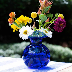 The VIETRI HIBISCUS BUD VASE COBALT is mouthblown with a translucent design, featuring a wide, flared wavy rim and bulbous base. Swirls of cobalt blue and purple elegantly blend throughout, creating an elegant and smooth appearance.