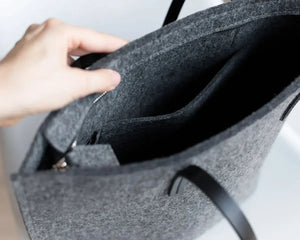 A person in a beige trench coat holds the WOOLBERRY WOOL FELT LONG TOTE BAG with black handles, standing against a plain, light-colored background.