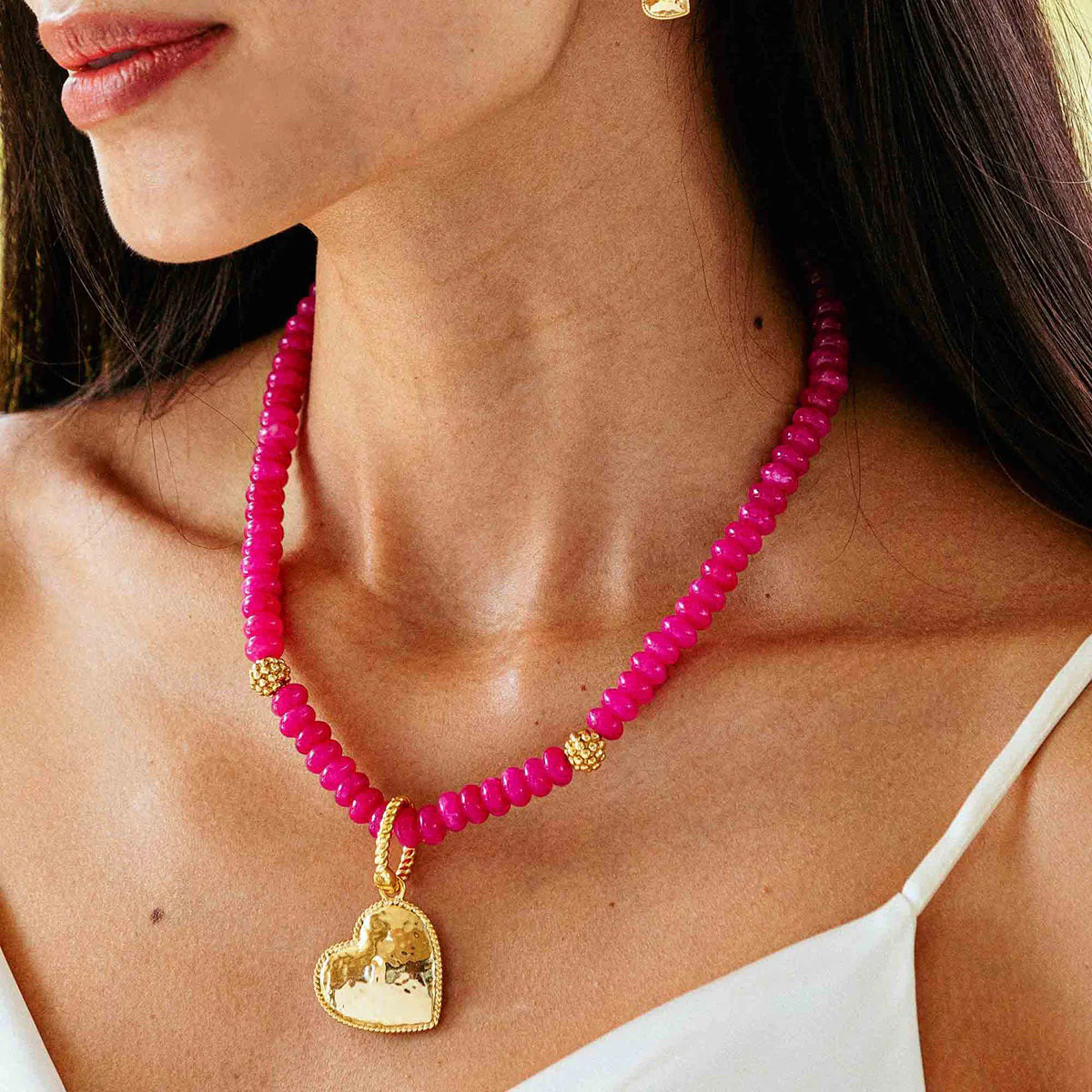 A woman wears the CAPUCINE DE WULF BERRY SINGLE STRAND NECKLACE, a vibrant pink bead necklace with gold accents and a large gold heart pendant. She has long dark hair and is dressed in a white spaghetti strap top. Her matching golden berries heart-shaped earring is visible, complementing the necklace perfectly.