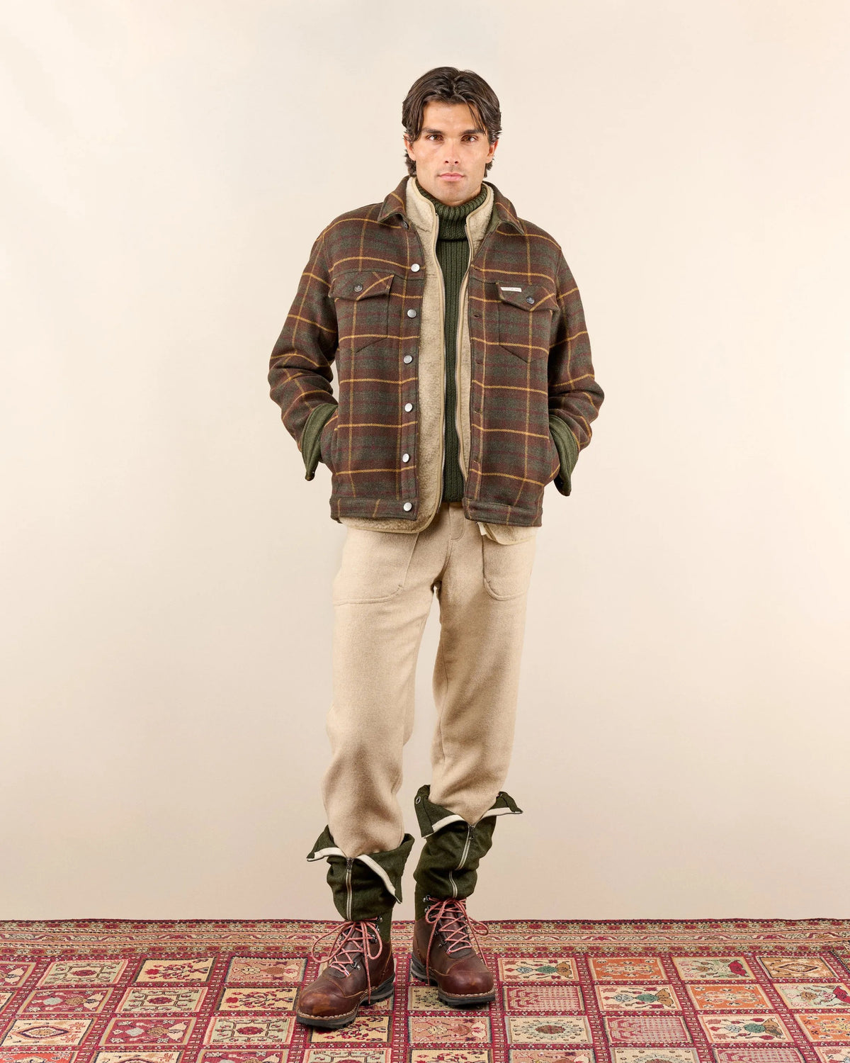 The AMUNDSEN SPORTS Wool Jacket is a unisex dark plaid overshirt with a brown and mustard check pattern, long sleeves, buttoned cuffs, and a small red, white, and blue striped tag near the back collar. Laid flat, viewed from behind.