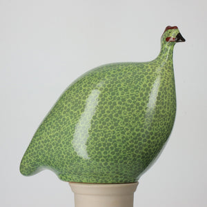 A handmade ceramic sculpture from LES CERAMIQUES DE LUSSAN, the LARGE CERAMIC FRENCH GUINEA in green spotted yellow, features a bird with a patterned body and small red accents on its head. Reminiscent of charming French partridges or quails, the bird is perched on a plain, light-colored cylindrical base against a simple white background.