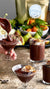 A hand is seen pouring the rich MAISON ZOE FORD - LUXURIOUS CHOCOLATE PUDDING MIX into a martini glass. Some of the glasses filled with this decadent chocolate pudding are topped with whipped cream and sprinkled nuts placed beside. In the background, there's a platter of fresh fruits and greens, adding a vibrant touch to the vegan dessert setup using oat milk from MAISON ZOE FORD.