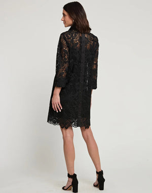 A person stands against a plain background wearing the Aileen Black Lace Dress by Hinson Wu, featuring knee-length black lace with 3/4 sleeves and button details. Paired elegantly with black high-heeled sandals, this outfit exudes a formal style perfect for the holiday season.