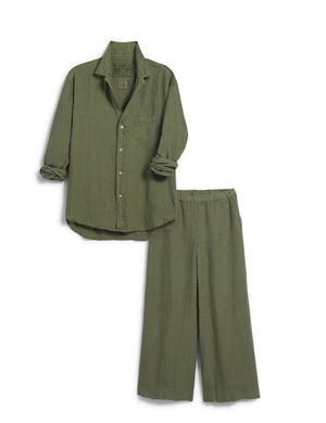 A person stands outdoors wearing an olive green FRANK AND EILEEN - EILEEN Relaxed Button Up Shirt in Linen from the Tuscany Getaway Set with the sleeves rolled to the forearms and matching Maisie pull-on pants. Their left hand is partially in their pocket, and greenery is visible in the background.