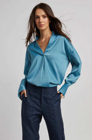 A person with long brown hair is standing with their back partially turned, wearing the ADROITE ATELIER - KAY POPOVER SATIN BLOUSE, an oversized white V-neck satin blouse from ADROIT ATELIER, paired with light blue jeans. The person is also wearing large, ornate earrings. The background is plain and light-colored.