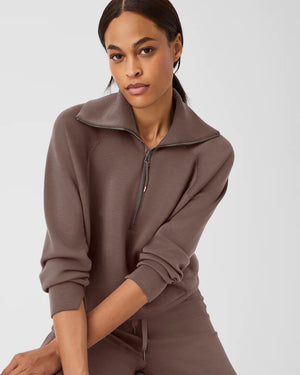 A woman with long brown hair is wearing the SPANX AIRESSENTIALS HALF ZIP sweatshirt and matching pants, both crafted from lightweight fabric for luxurious comfort. She holds her braided ponytail with one hand while looking to her right against a plain white background.