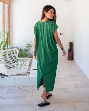 A woman stands in a well-lit hallway, wearing the vibrant MERSEA - IBIZA KAFTAN DRESS, accessorized with a necklace and black sandals. The lightweight green dress is complemented by a white chair and greenery in the background, creating a resort-worthy, modern atmosphere.