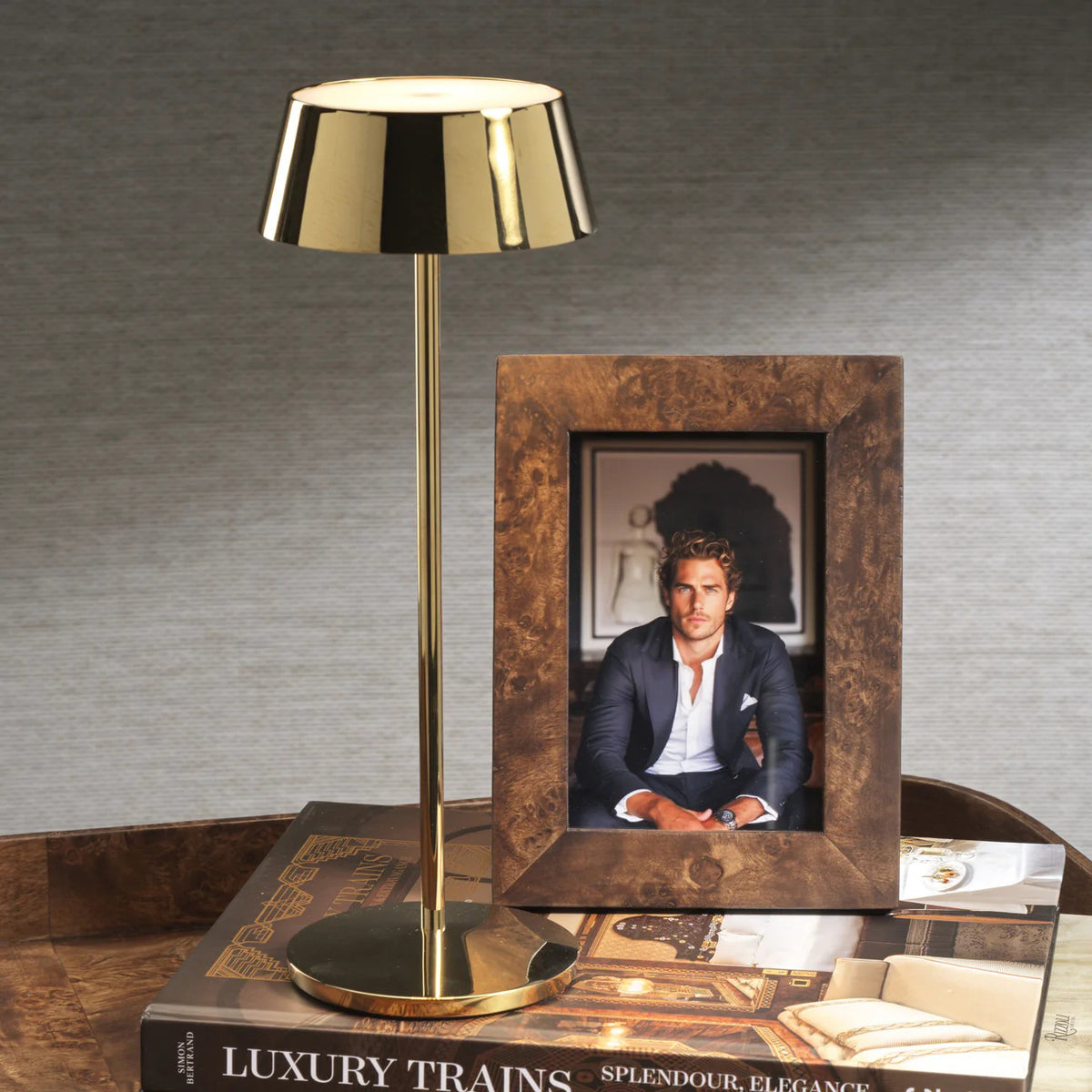 A gold table lamp stands beside the ZODAX BURL WOOD PHOTO FRAME 4X6 displaying a photo of a man in a suit, both set atop large books—including &quot;Luxury Trains&quot;—on a wooden surface with a gray backdrop.