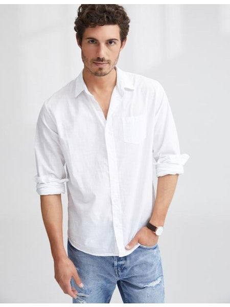A man with short curly hair wears the FRANK &amp; EILEEN LUKE Men&#39;s Button Up Shirt in Classic White Poplin (sleeves rolled), paired with light blue ripped jeans and a watch, smiling slightly against a plain background.