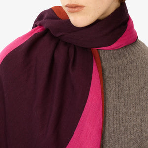 Close-up of a person wearing the INOUI EDITIONS - AMOUR SCARF in purple and pink cashmere, wrapped around their neck. The person has a neutral expression and is dressed in a grey sweater. The plain white background highlights the winter accessory from INOUI and part of the sweater.