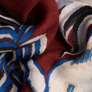 A person is wearing a cream-colored sweater with the INOUI EDITIONS - WESTERN SQUARE SCARF from INOUI, a large, burgundy wool scarf adorned with unique prints draped around their neck. The image focuses on the upper body, showcasing the intricate design of the scarf against a plain white background.