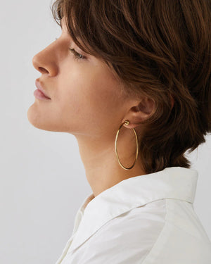 Two elegant Icon Hoop Earrings Medium by JENNYBIRD in high-polish gold are displayed against a light background. Each earring features a small spherical closure. One lies flat while the other stands upright, highlighting their minimalistic design crafted from stainless steel.