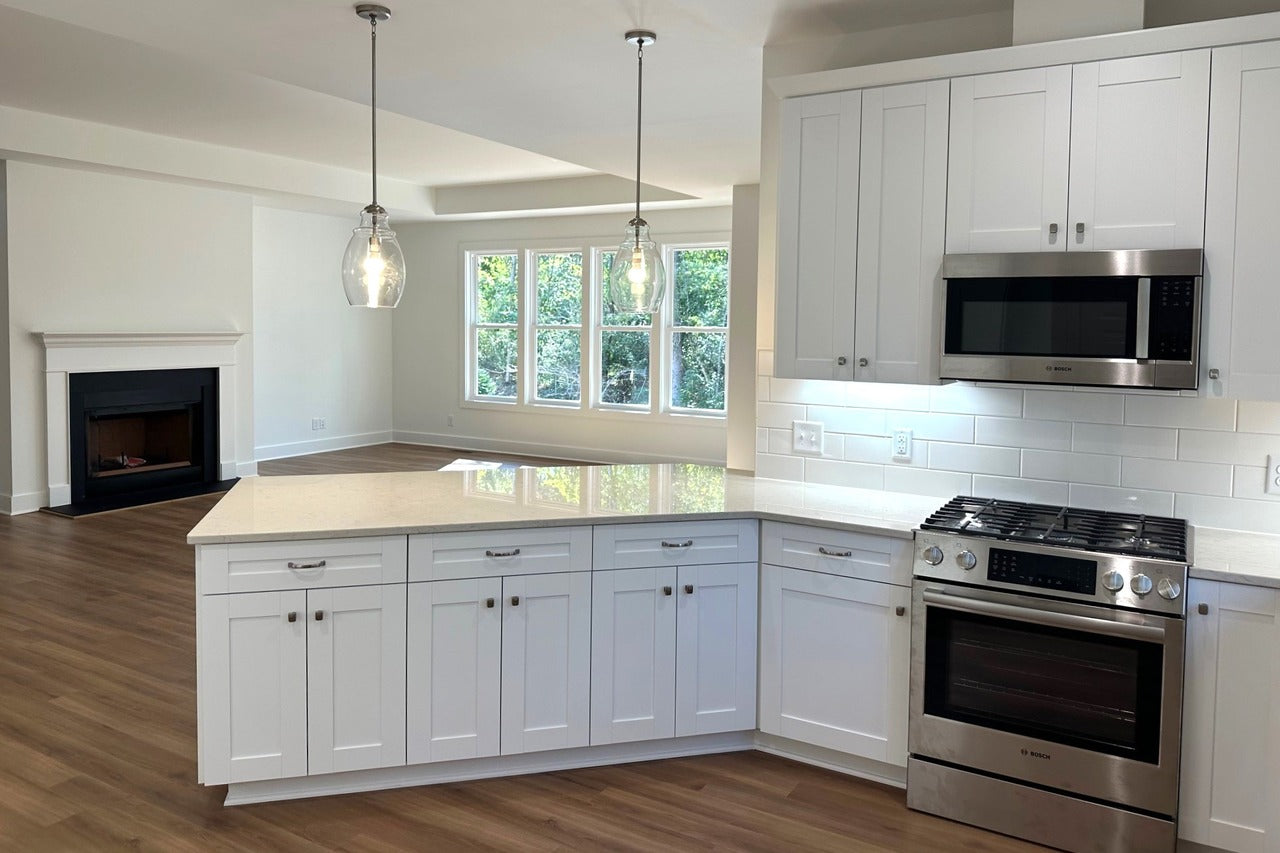 Modern kitchen with white cabinets, quartz countertops, stainless steel appliances, and pendant lights. Large windows and wood floors, with a view of a living area featuring a fireplace. Bright, open, and airy space.