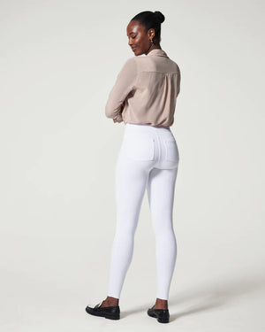 A person stands confidently in front of a plain background. They are wearing a white sleeveless turtleneck top, SPANX - JEANNISH ANKLE LEGGINGS in dark blue, and black and white loafers. Their arms are relaxed, and they gaze directly into the camera.