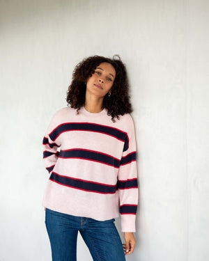 A person with curly hair stands against a light wall, wearing the ultra-soft MERSEA - KEEPER CREWNECK SWEATER in light pink with black and red stripes, paired with blue jeans. The individual faces away, looking to the left.